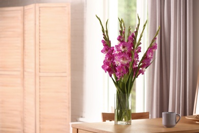 Vase with beautiful pink gladiolus flowers, pictures and cup on wooden table in room, space for text