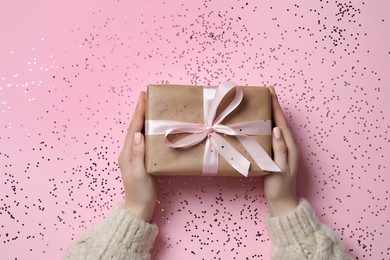 Photo of Christmas present. Woman holding gift box and confetti on pink background, top view