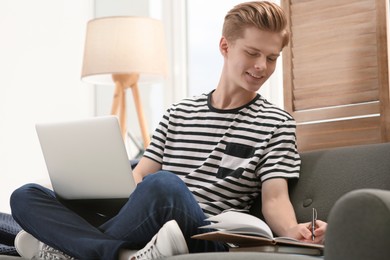 Online learning. Smiling teenage boy with laptop writing in notebook on sofa at home