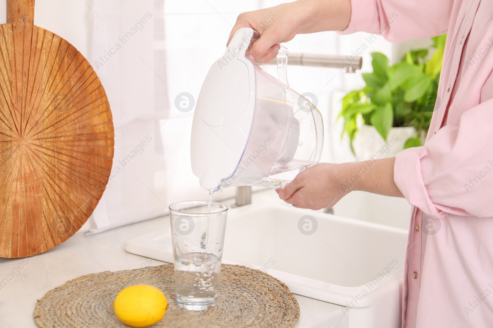 Photo of Woman pouring water from filter jug into glass in kitchen, closeup