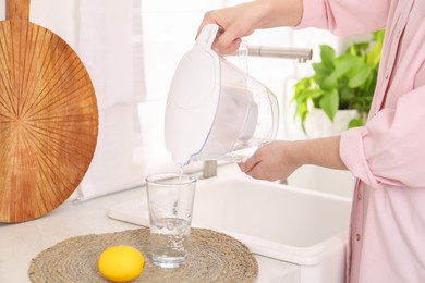 Photo of Woman pouring water from filter jug into glass in kitchen, closeup