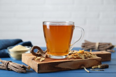 Photo of Aromatic licorice tea in cup and dried sticks of licorice root on blue wooden table