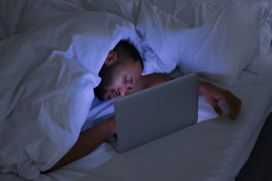 Photo of Young man sleeping near laptop under blanket in bed at night. Internet addiction