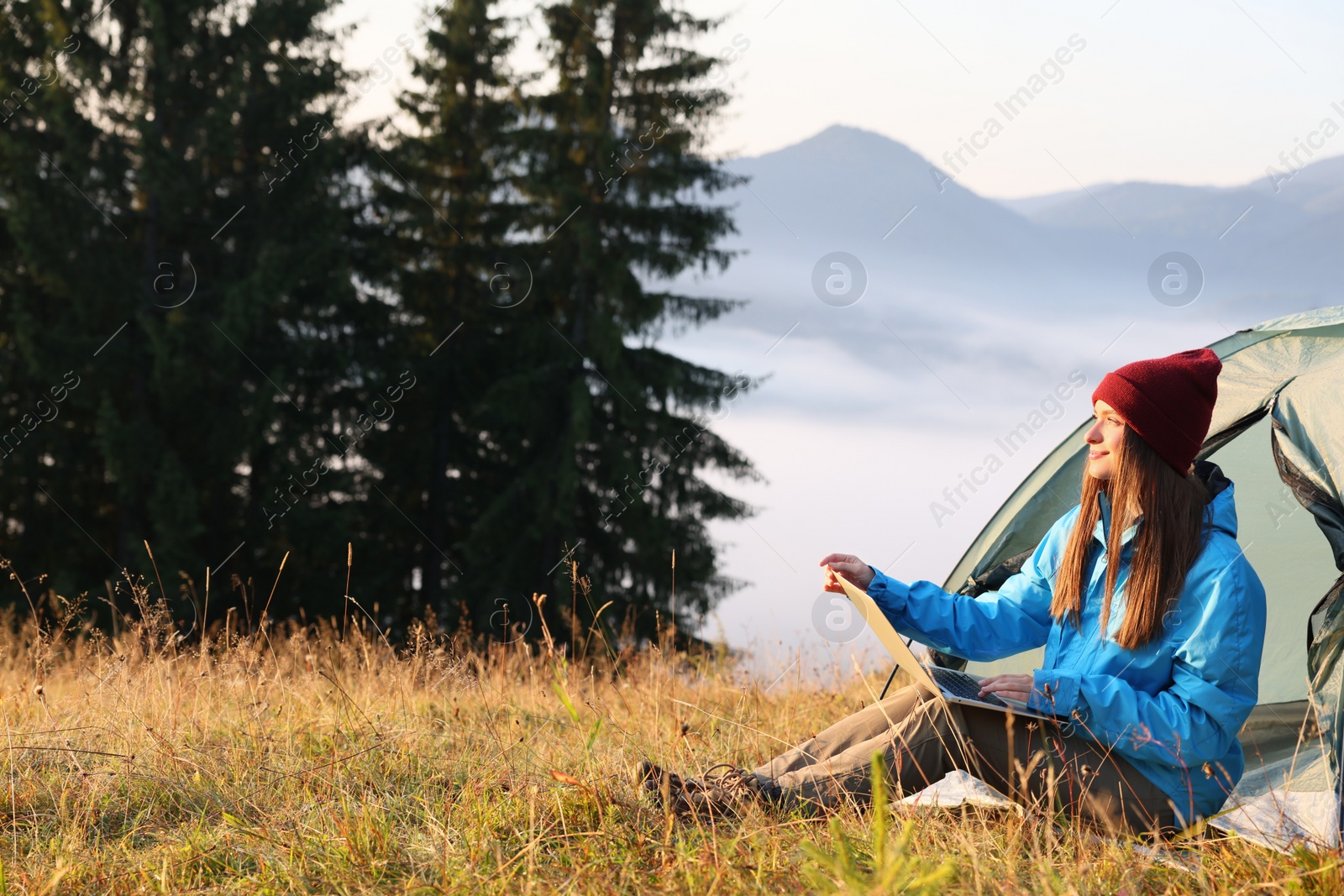 Photo of Woman working on laptop near camping tent outdoors surrounded by beautiful nature