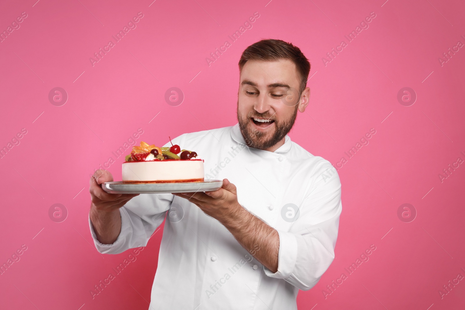 Photo of Happy professional confectioner in uniform holding delicious cake on pink background