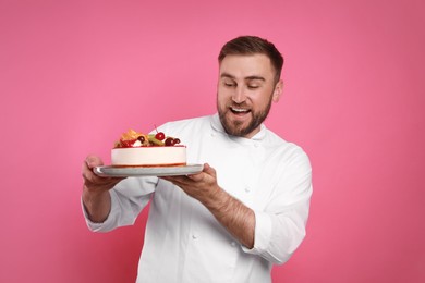 Happy professional confectioner in uniform holding delicious cake on pink background