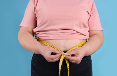 Photo of Overweight woman measuring waist with tape on light blue background, closeup