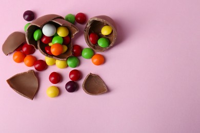 Photo of Broken chocolate egg with colorful candies on pink background, flat lay. Space for text