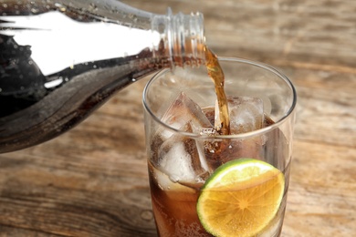 Photo of Pouring refreshing soda drink into glass with ice cubes and lime on wooden table, closeup