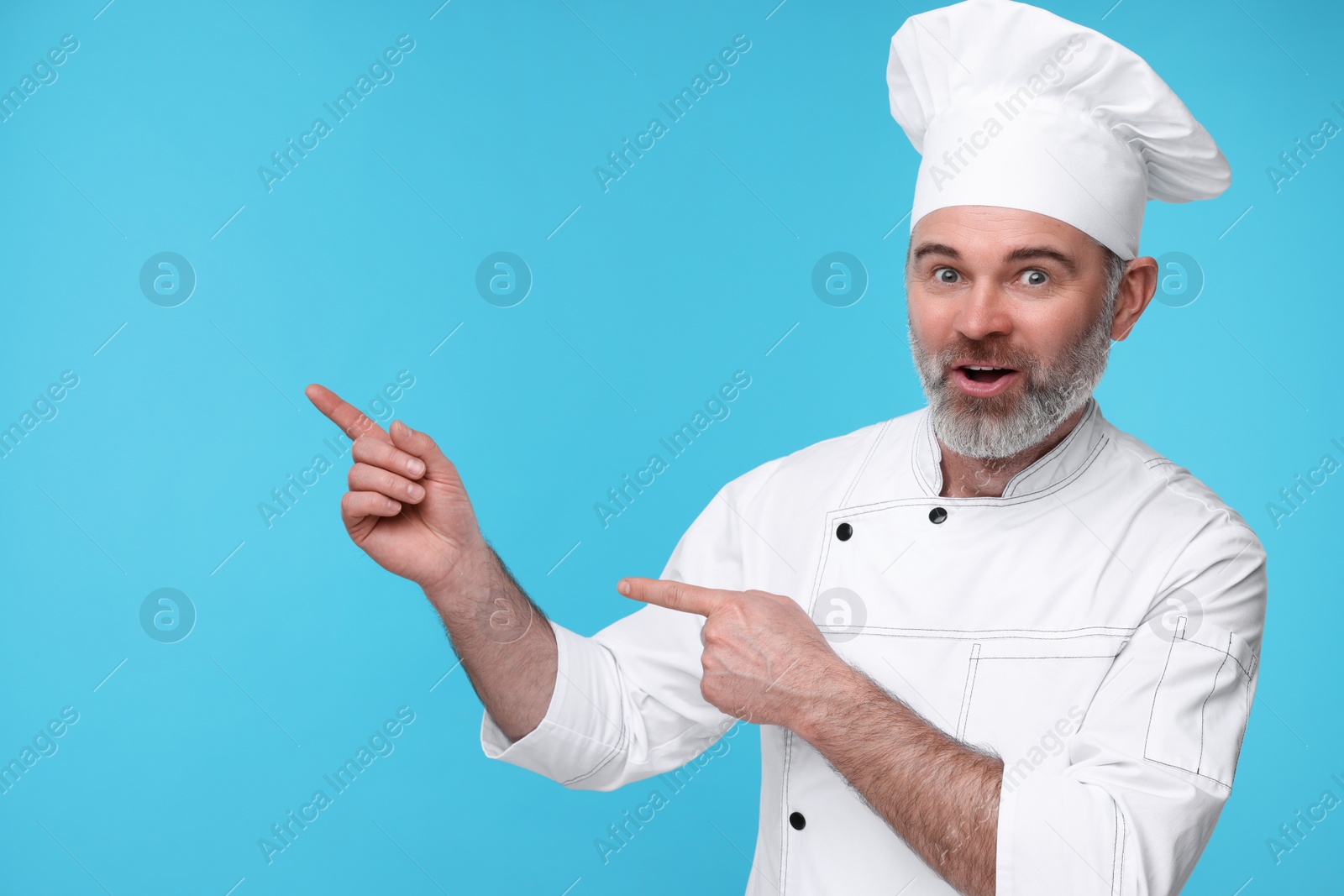 Photo of Surprised chef in uniform pointing at something on light blue background