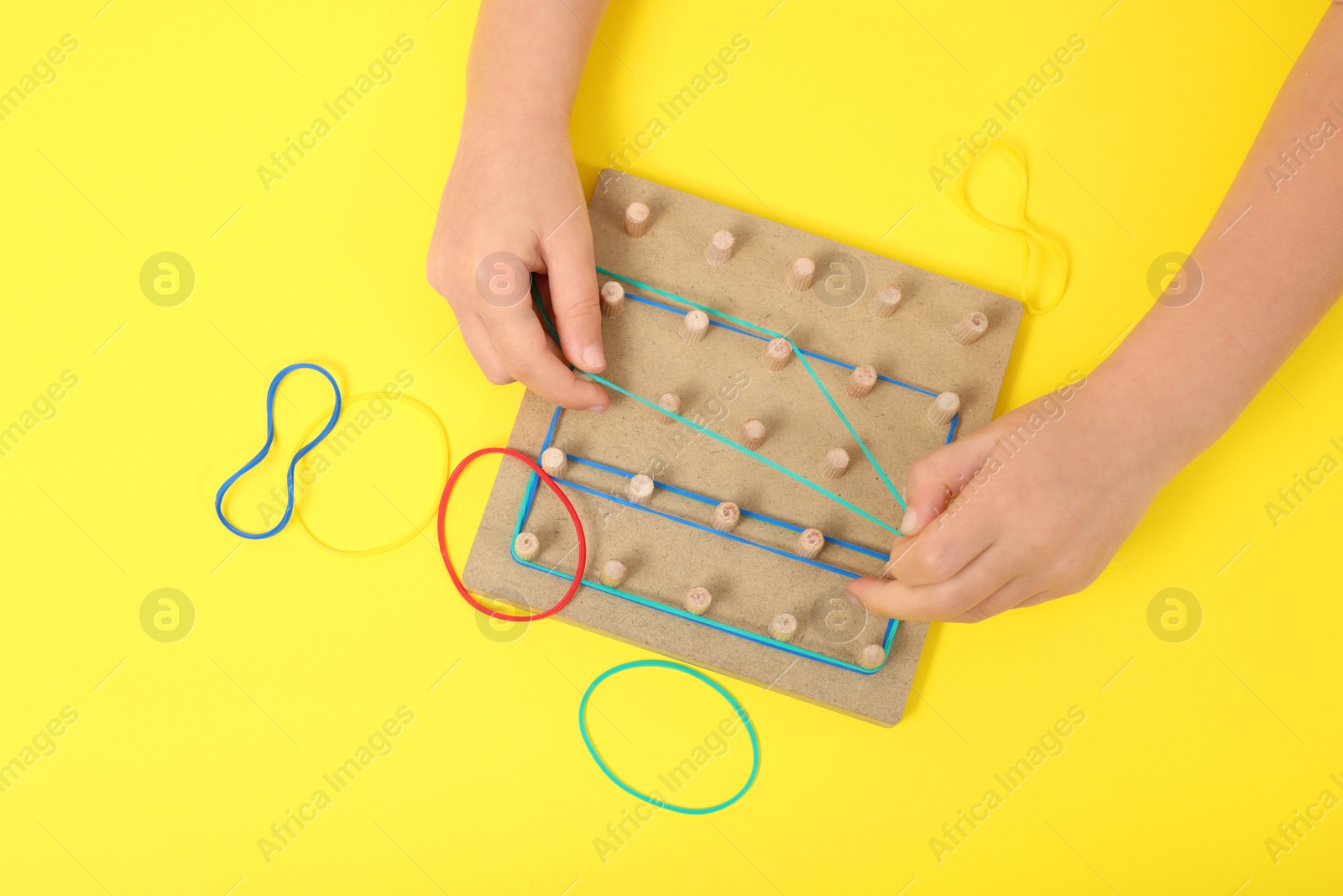 Photo of Motor skills development. Girl playing with geoboard and rubber bands at yellow table, top view