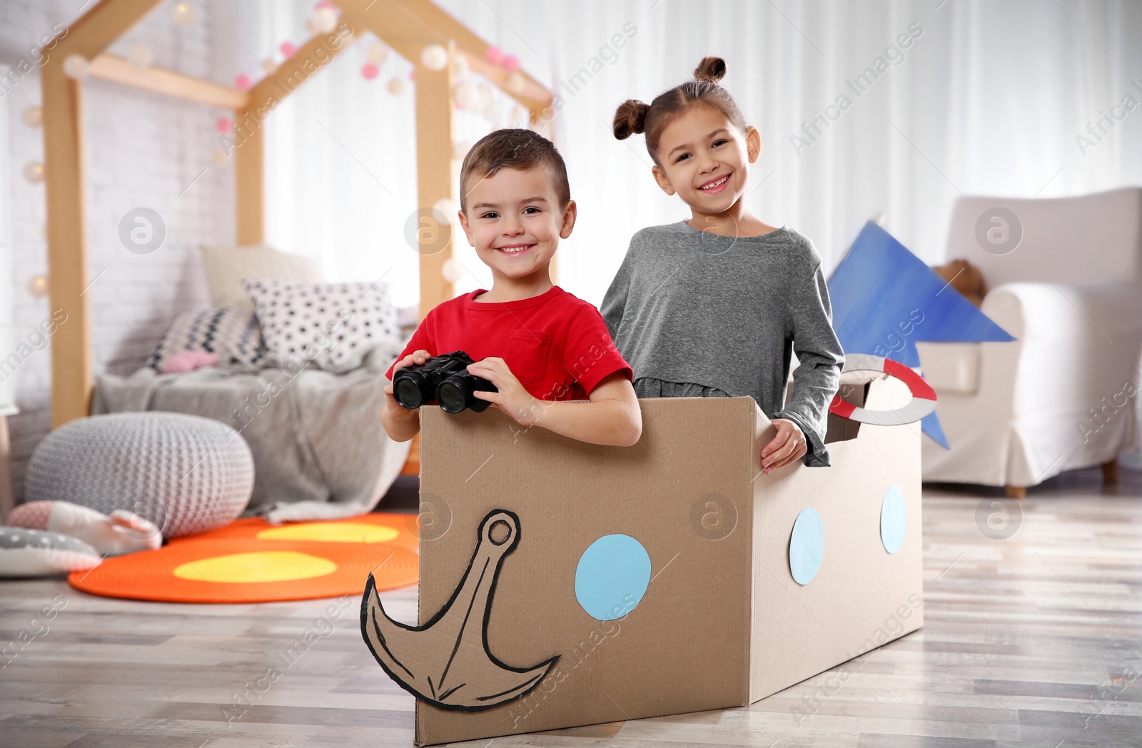 Photo of Cute little kids playing with binoculars and cardboard boat in bedroom