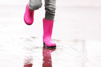 Photo of Little girl wearing rubber boots splashing in puddle on rainy day, focus of legs. Autumn walk