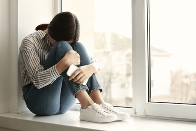 Photo of Young woman with mobile phone crying on window sill, space for text. Loneliness concept