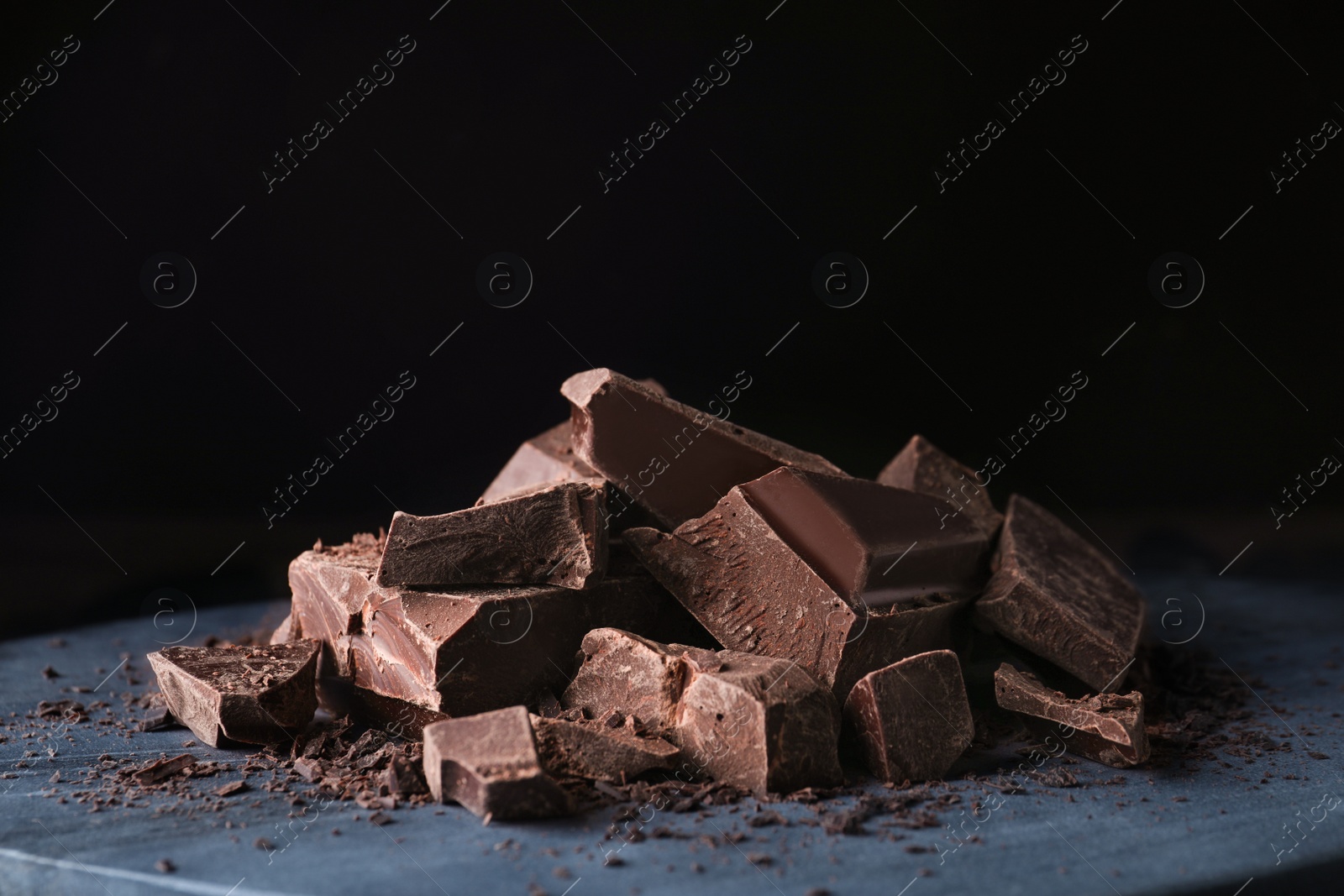 Photo of Pieces of tasty dark chocolate on grey board against black background