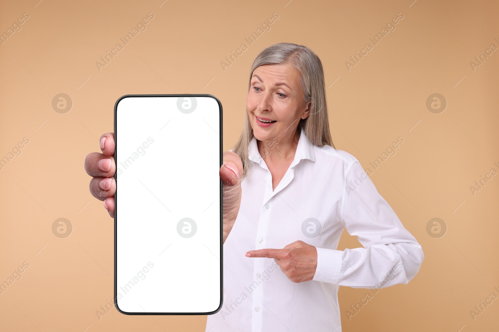 Image of Happy mature woman pointing at mobile phone with blank screen on beige background. Mockup for design