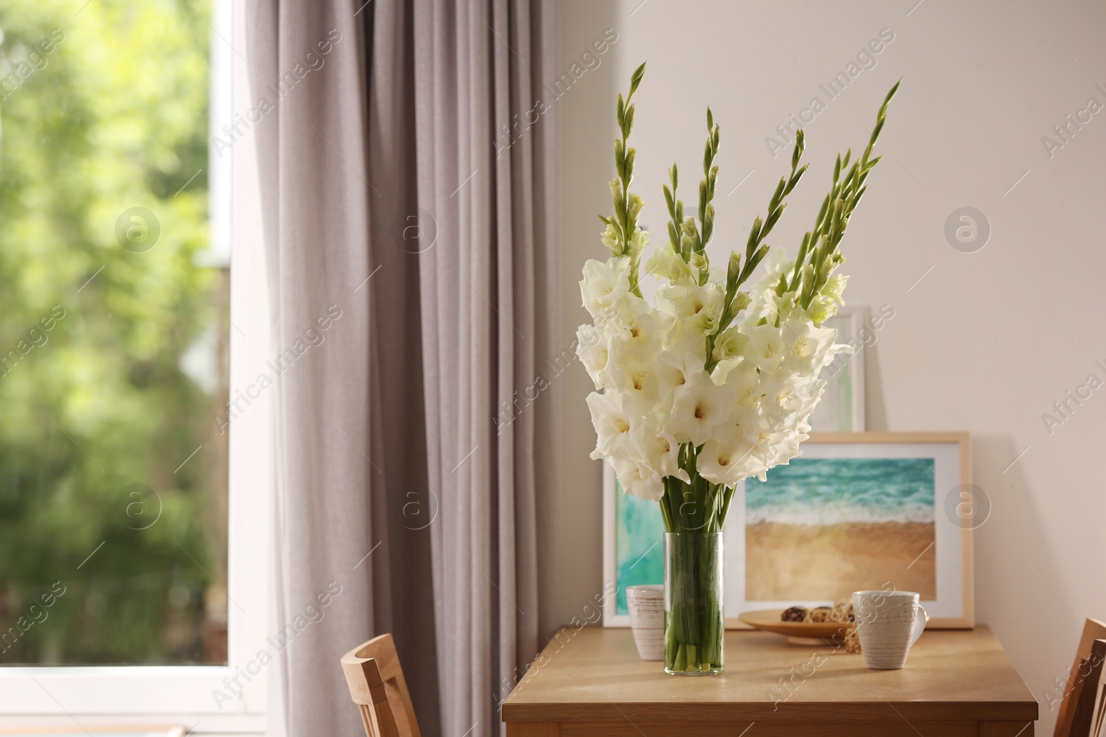 Photo of Vase with beautiful white gladiolus flowers, pictures and cups on wooden table in room, space for text