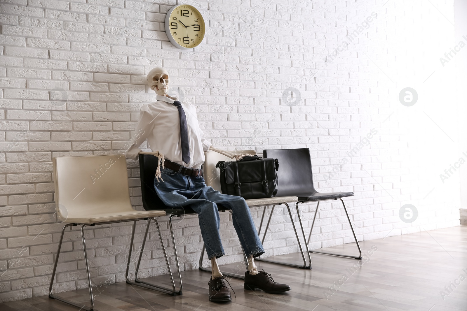 Photo of Human skeleton in office wear sitting on chair near brick wall indoors