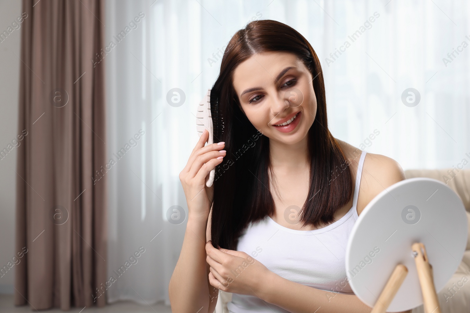 Photo of Beautiful woman brushing her hair near mirror in room, space for text