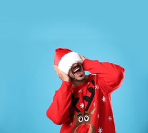 Photo of Emotional young man in Christmas sweater and Santa hat on light blue background