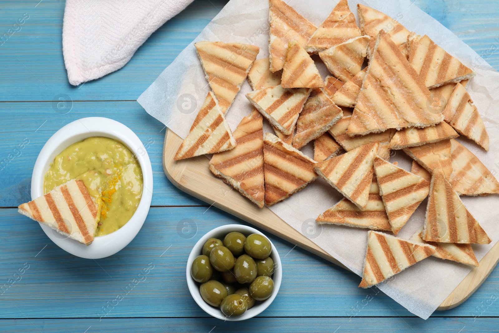 Photo of Delicious pita chips, olives and hummus on light blue wooden table, flat lay