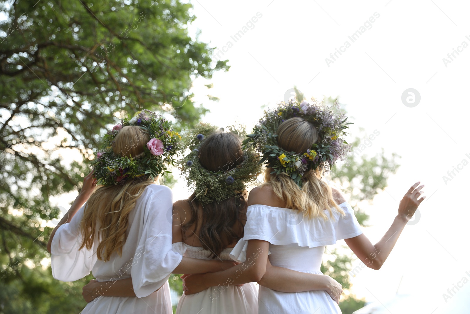 Photo of Young women wearing wreaths made of beautiful flowers outdoors on sunny day, back view