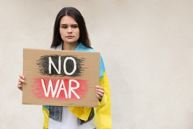Photo of Sad woman with Ukrainian flag holding poster with words No War against light wall. Space for text