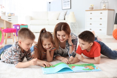 Photo of Cute little children reading book on floor with young mother in playing room