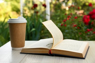 Photo of Open book with paper cup of coffee on wooden table in garden