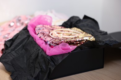 Photo of Headbands and stylish pink carnival costume with sequins in black box on wooden table, closeup