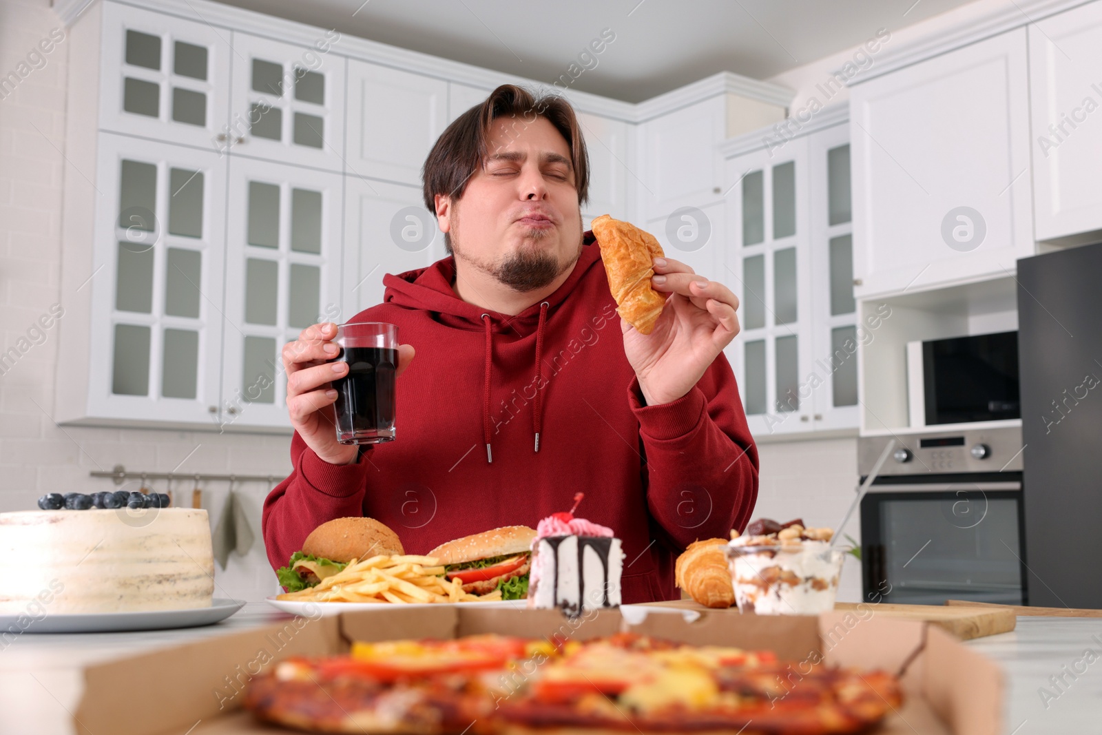 Photo of Overweight man eating tasty croissant at table in kitchen