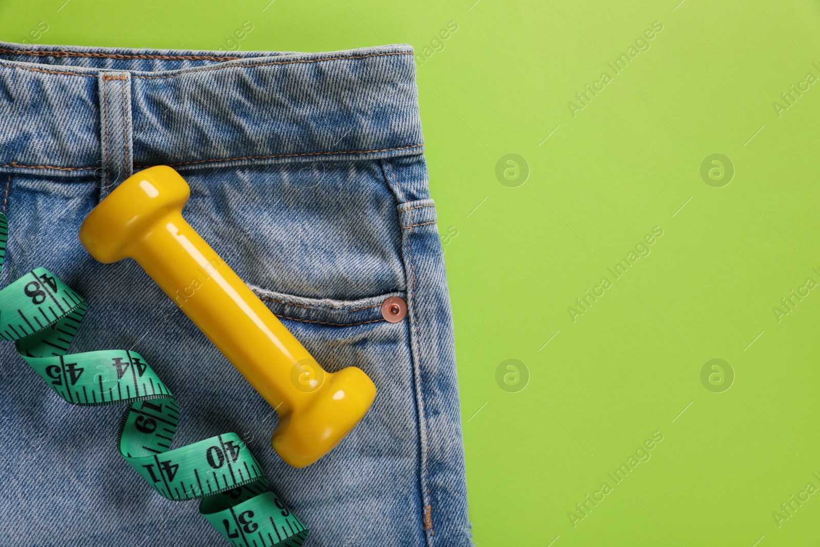Photo of Jeans, dumbbell and measuring tape on light green background, top view. Weight loss concept