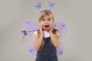 Photo of Surprised little girl in fairy costume with violet wings and magic wand on light grey background
