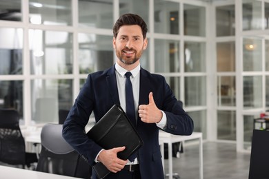 Photo of Happy real estate agent with leather portfolio showing thumb up indoors