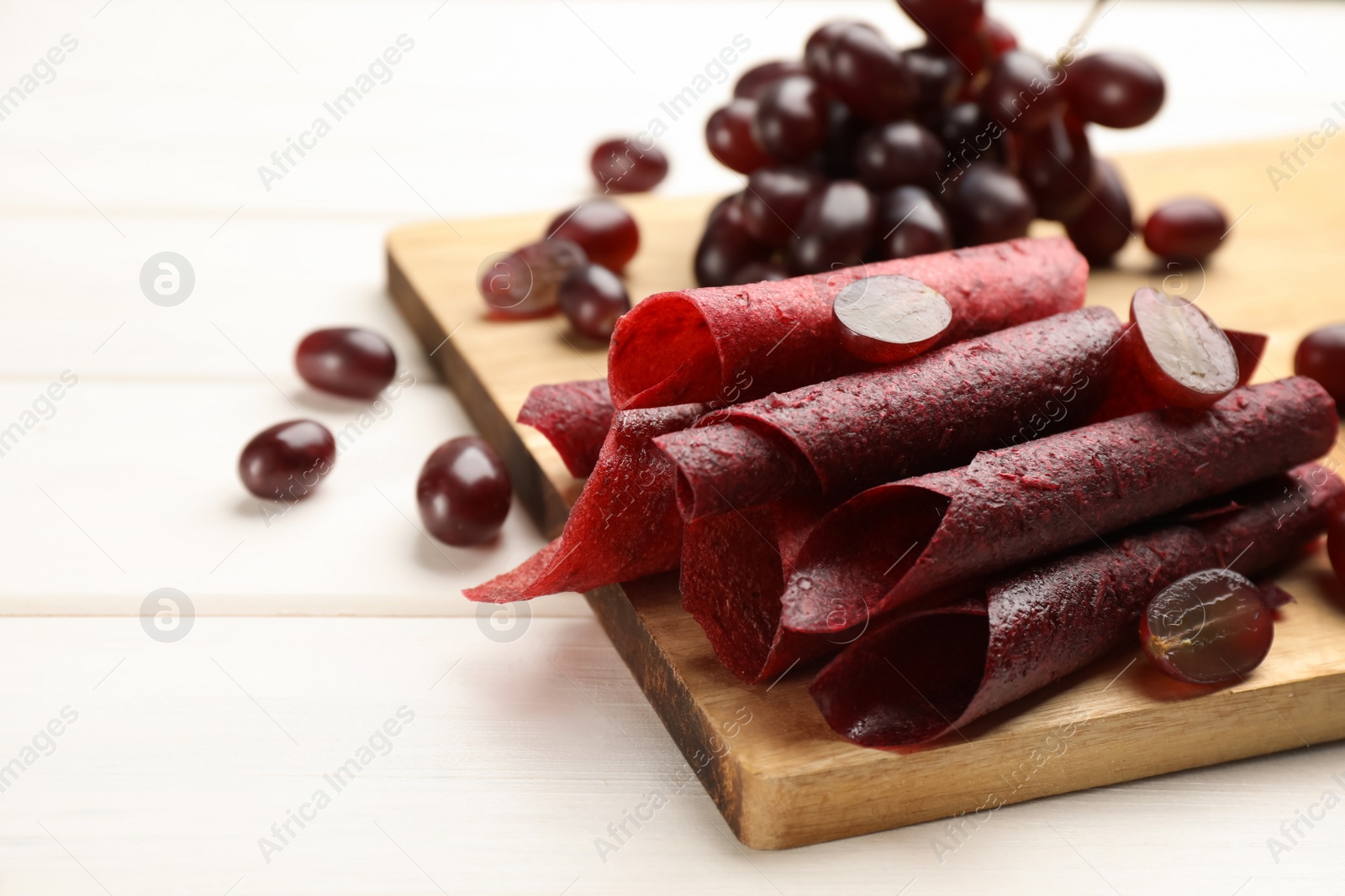 Photo of Delicious fruit leather rolls and grapes on white wooden table. Space for text