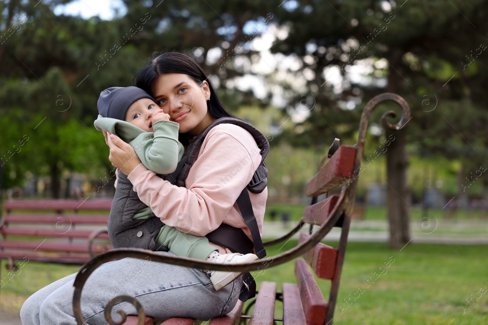 Photo of Mother holding her child in sling (baby carrier) on bench in park