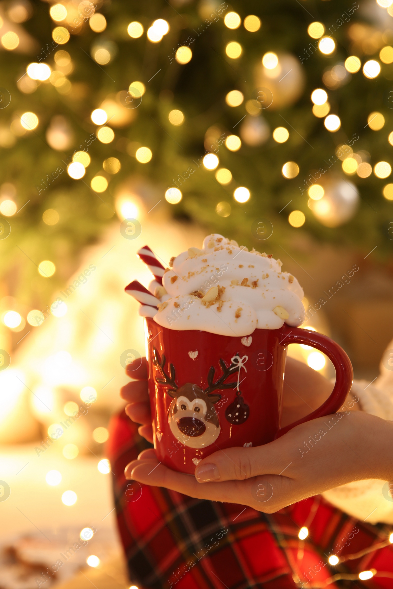 Photo of Woman holding cup of delicious drink with whipped cream near Christmas tree indoors, closeup