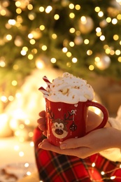 Photo of Woman holding cup of delicious drink with whipped cream near Christmas tree indoors, closeup
