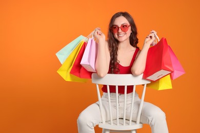 Happy woman in stylish sunglasses with many colorful shopping bags on chair against orange background