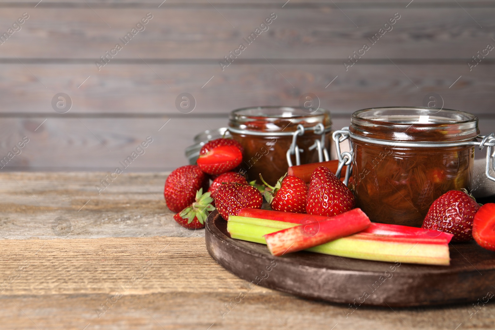 Photo of Jars of tasty rhubarb jam, fresh stems and strawberries on wooden table. Space for text