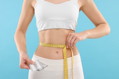 Photo of Slim woman holding pills and measuring waist with tape on light blue background, closeup. Weight loss