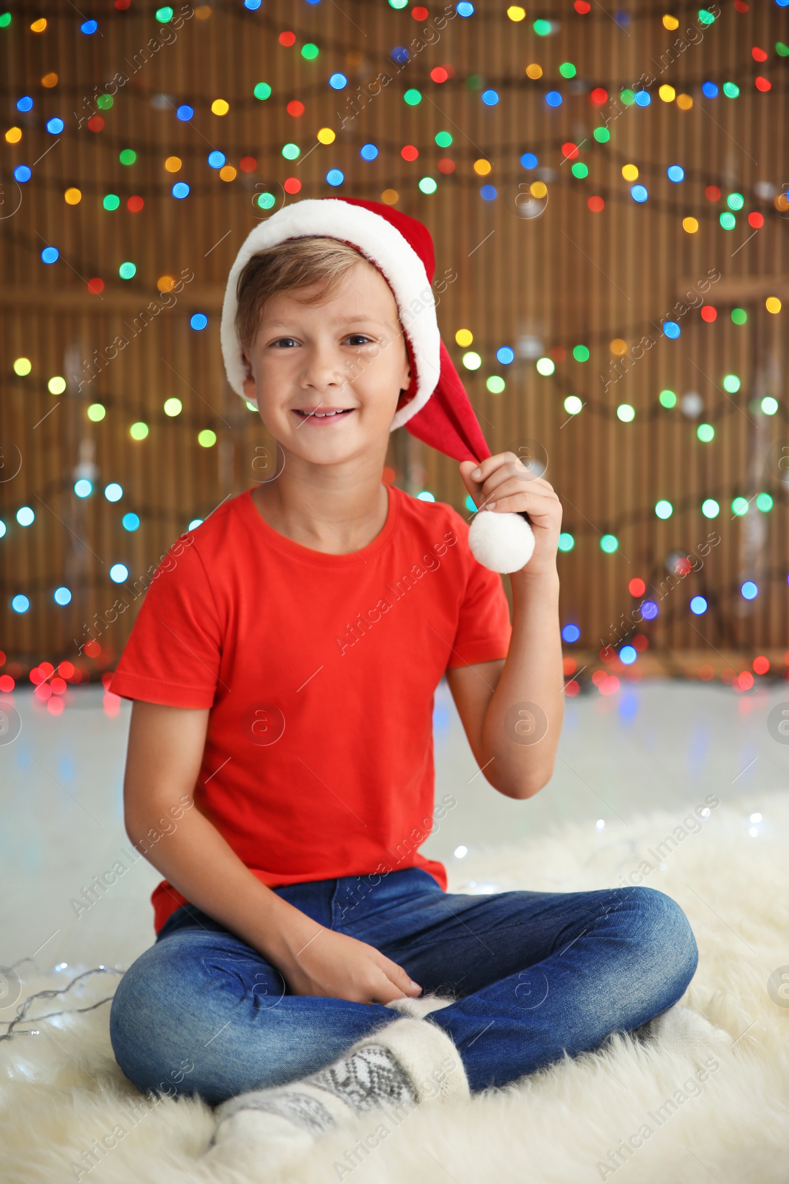 Photo of Cute little child in Santa hat on blurred lights background. Christmas celebration