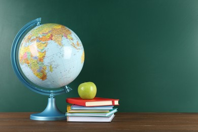 Globe, books and apple on wooden table near green chalkboard, space for text. Geography lesson
