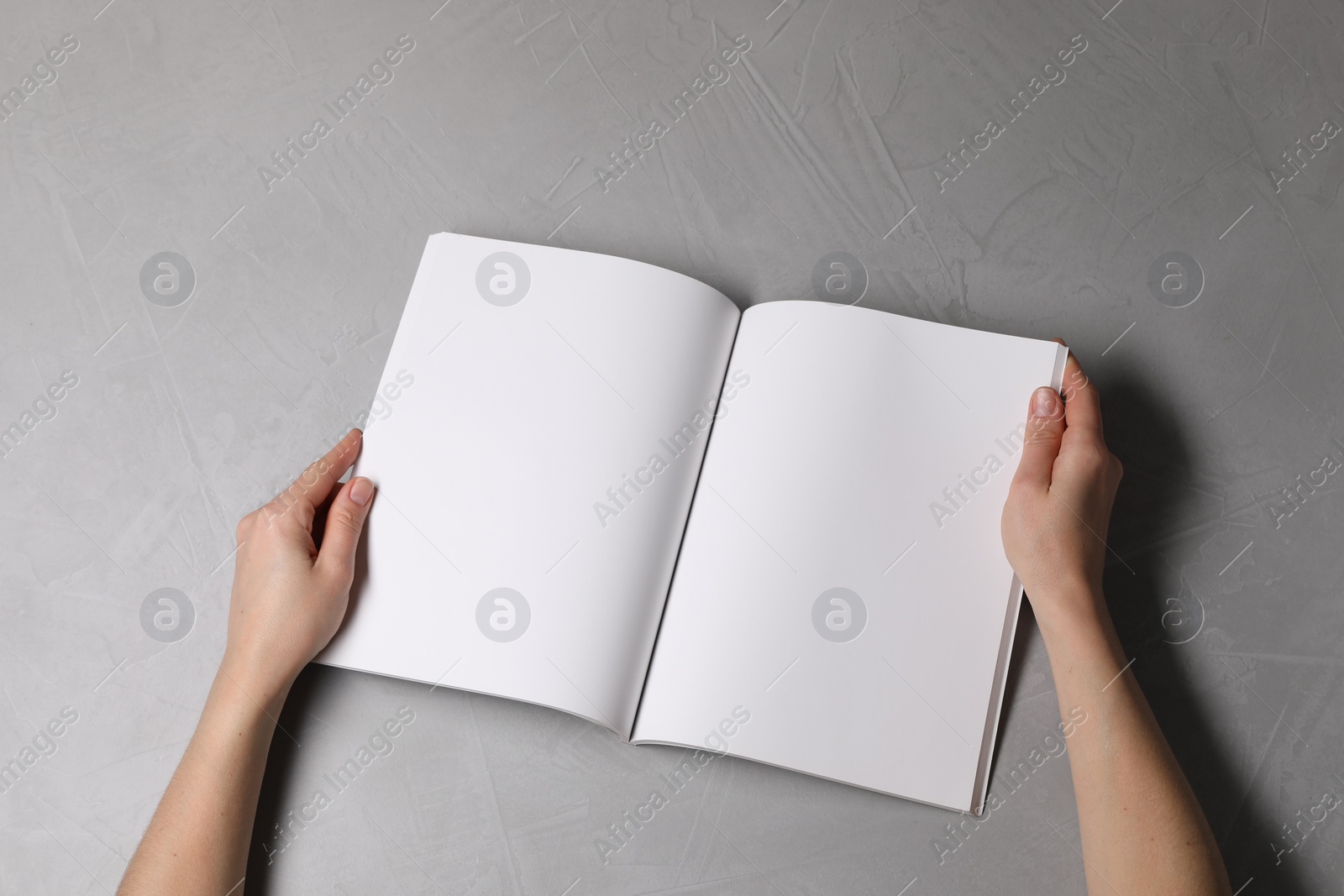 Photo of Woman holding open notebook with blank pages at light grey table, top view. Mockup for design