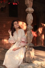 Beautiful woman in embroidered dress sitting near carved wooden column on terrace. Ukrainian national clothes