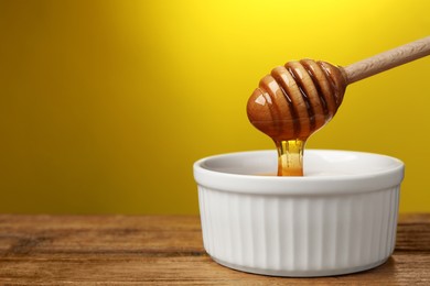 Photo of Pouring honey from dipper into bowl at wooden table against yellow background, closeup. Space for text