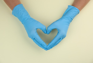 Person in medical gloves making heart with hands on beige background, top view
