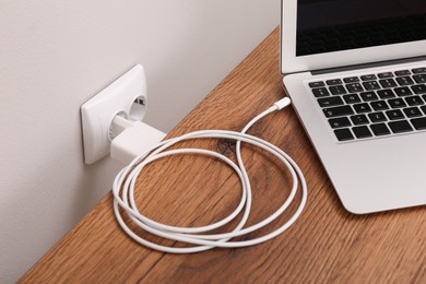Photo of Charging laptop with power adapter in electrical socket on wooden table