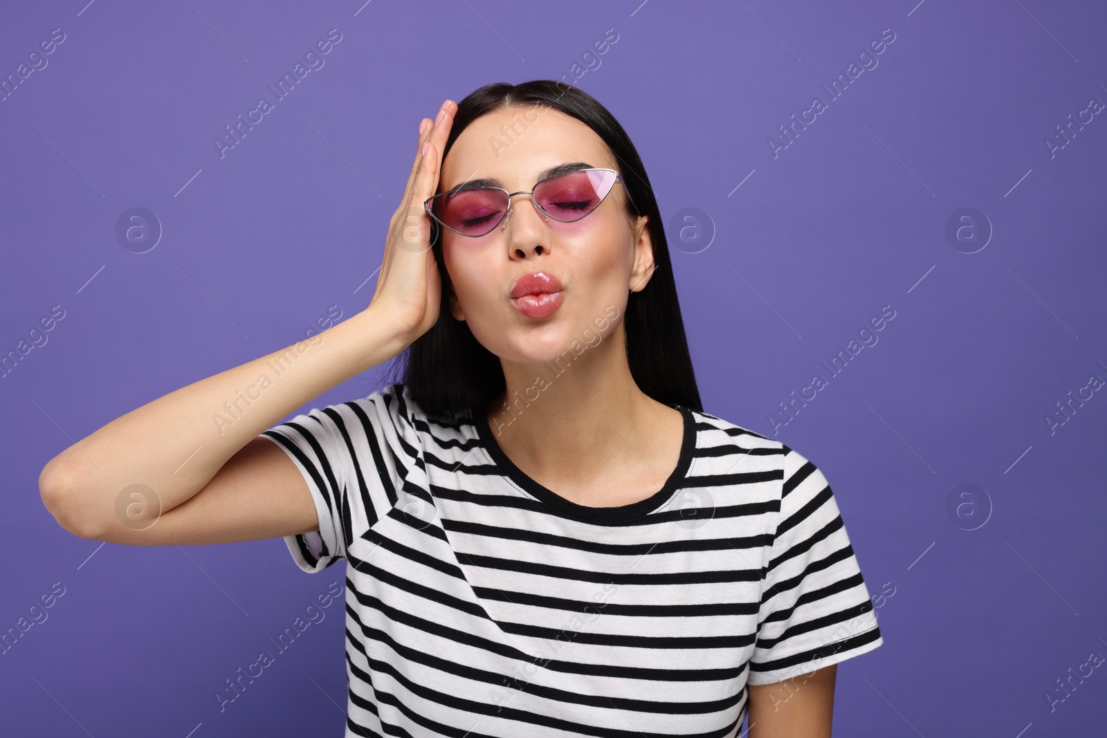 Photo of Beautiful young woman in stylish sunglasses blowing kiss on purple background