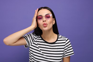 Beautiful young woman in stylish sunglasses blowing kiss on purple background